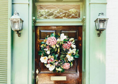 Carolina Electrical Supply Company | outdoor sconces of a light green house on either side of a dark wood door with leaded glass overhead and a pink and ivory floral wreath hanging on the door