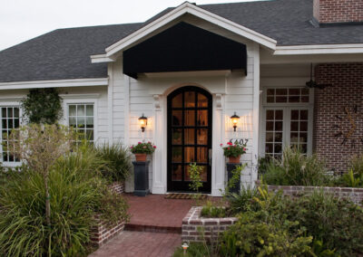 Carolina Electrical Supply Company | lit outdoor sconces by front door of a white craftsmen style home with a brick porch and walkway leading up to the house
